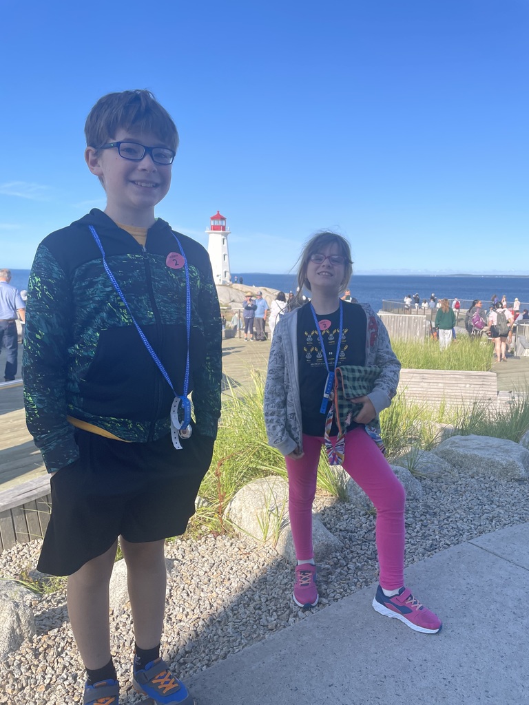 Chase and Abby stand on a bed of small rocks, with a white and red light house, water and tourists in the background