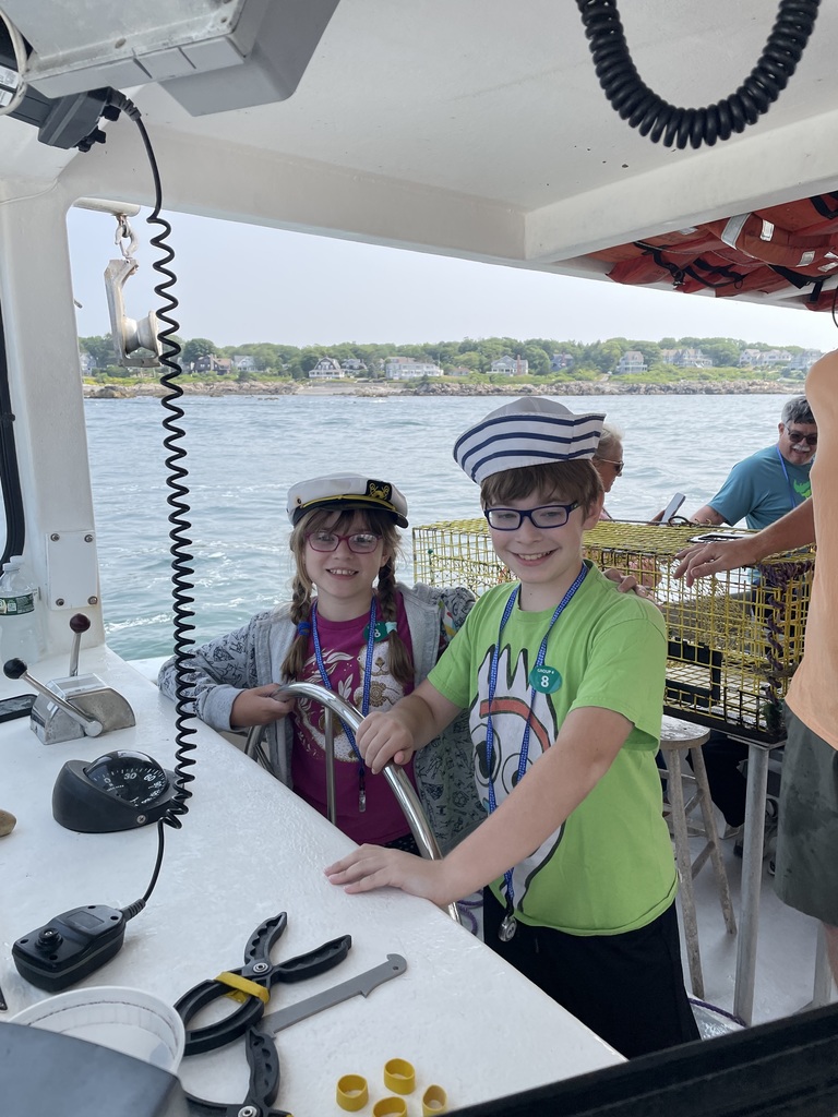 Chase and Abby stand on the deck of a boat, with Chase at the steering wheel, wearing sailor hats
