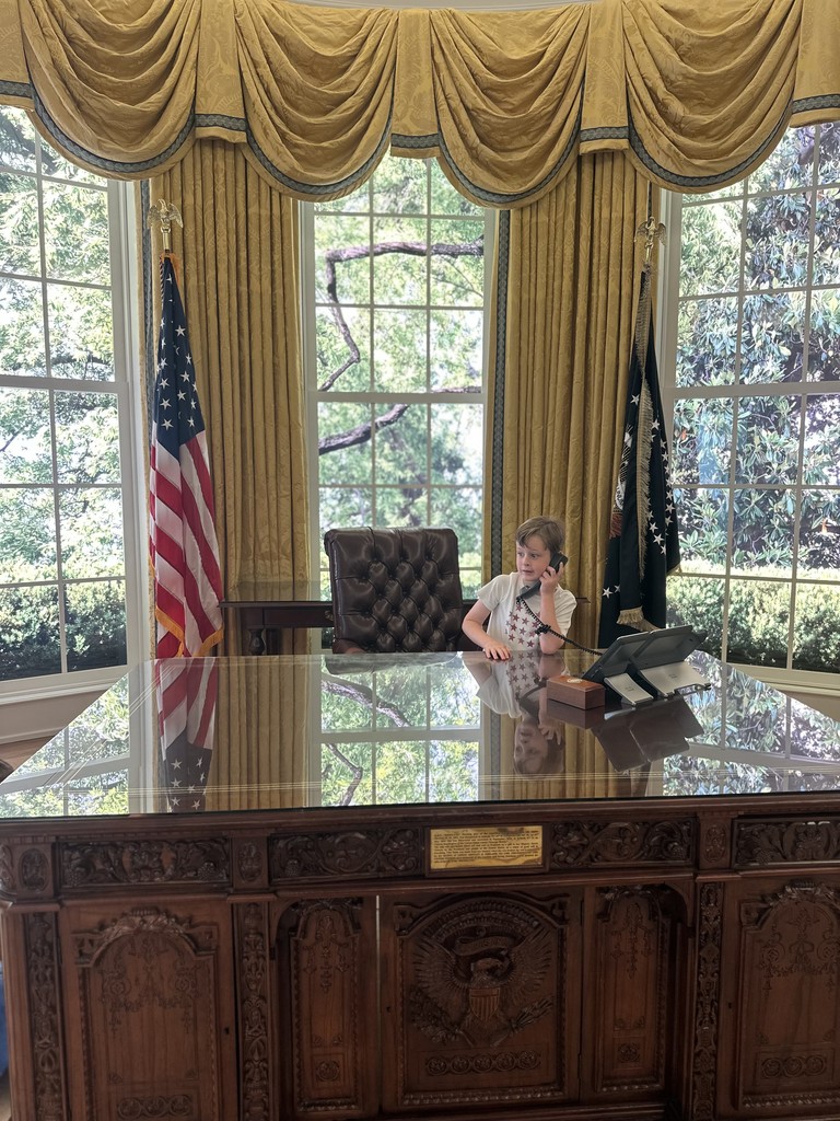 Robbie stands on the phone at a huge beautiful desk with an American flag, tall curtained windows and scenery behind him