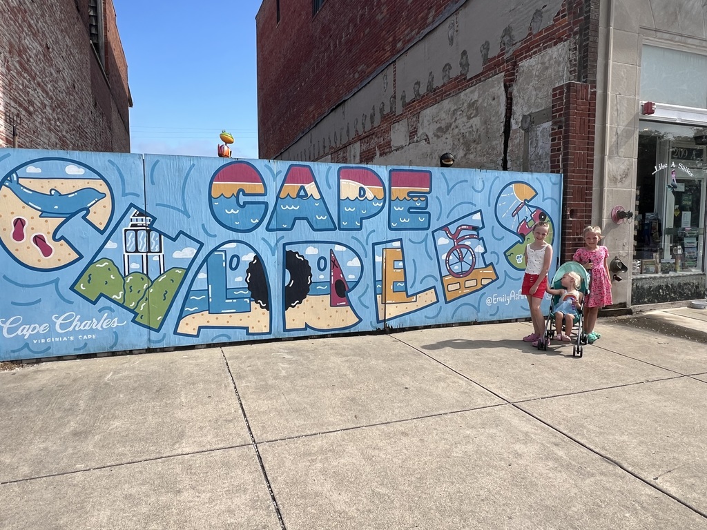 Sheileen and Annaleia, as well as a young sibling in a stroller, stand in front of a colorful mural on a sidewalk that says "Cape Charles"