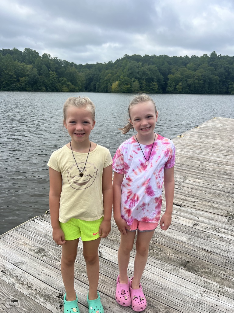 Sheileen and Annaleia are standing on a dock smiling with a body of water and a horizon line filled with trees in the background