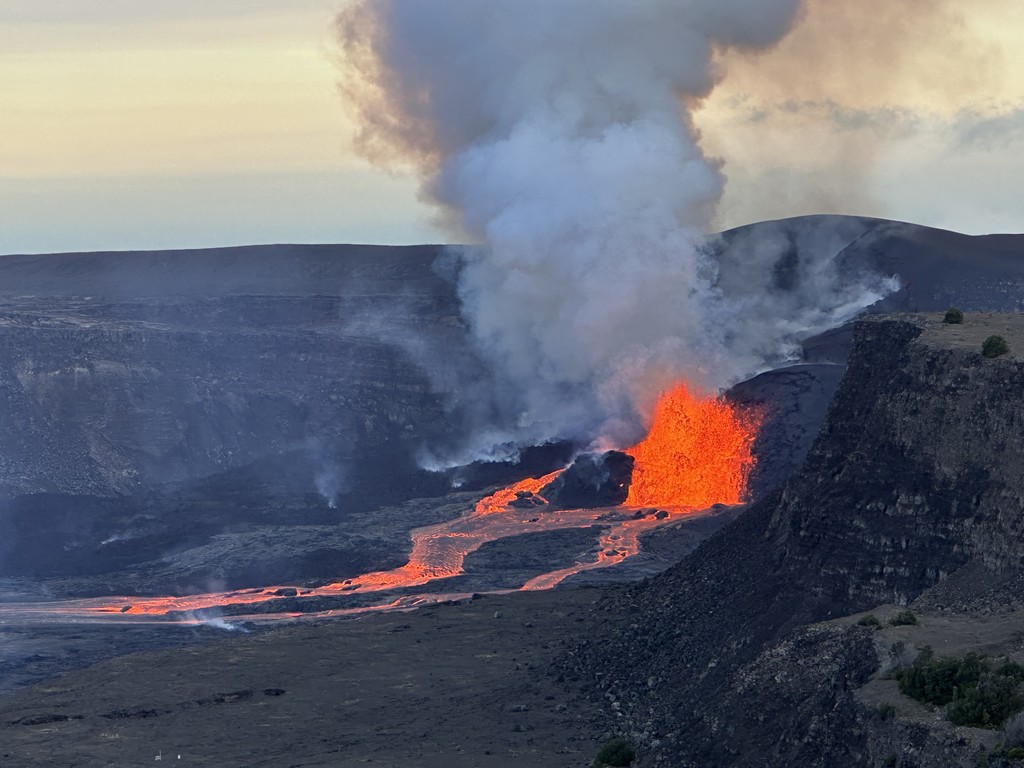 The Kilauea volcano erupting with lava spilling over the endges into the ground below and smoke billowing into the sky