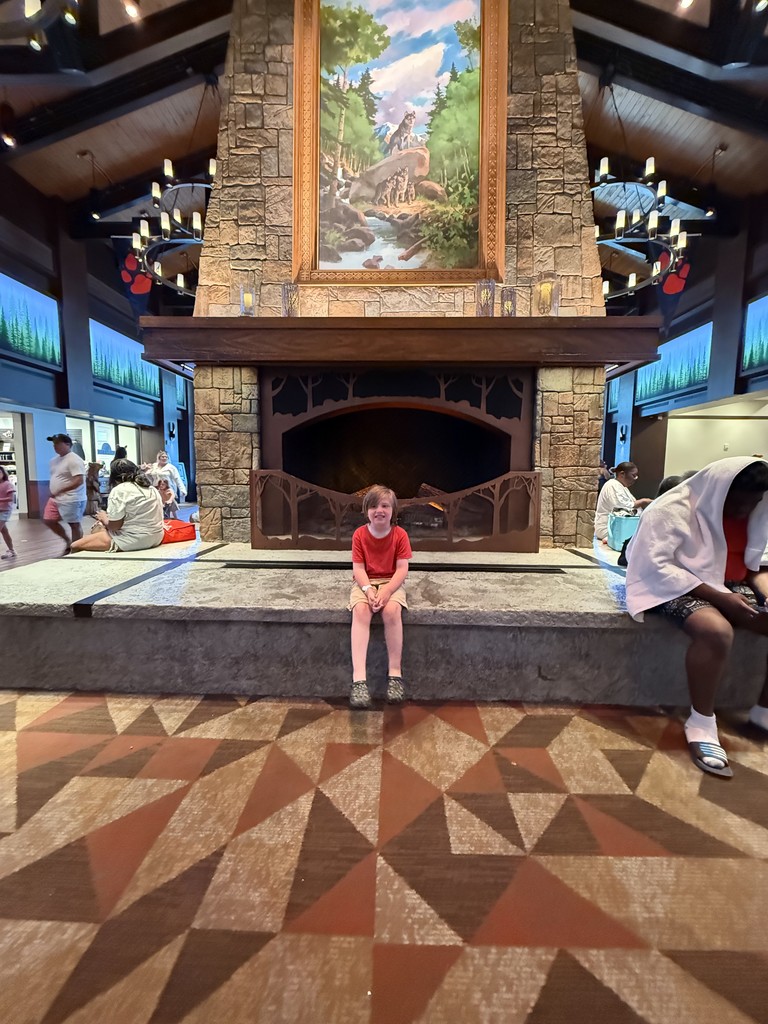 Richard sits at the base of a huge fireplace with a framed picture of wolves over the wooden mantel, with candelabra chandeliers overhead and the lodge lobby around him
