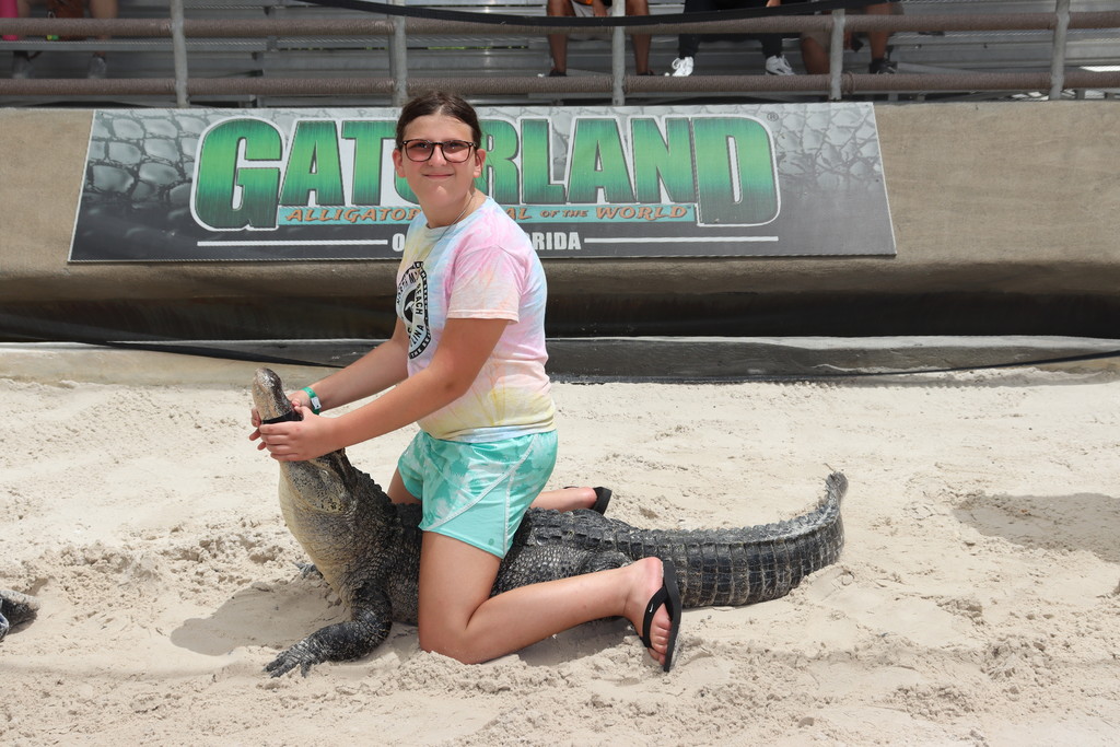 Ella sits on an alligator among sand with a "Gatorland" sign in the background