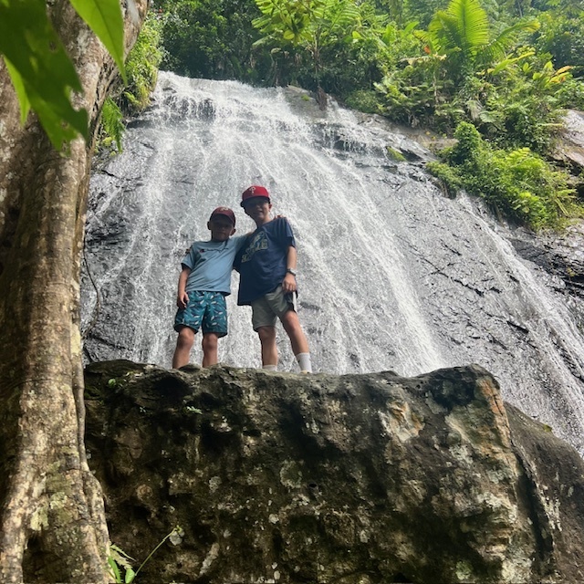 Jack & Francis stand on a rock cliff among trees with a waterfall behind them