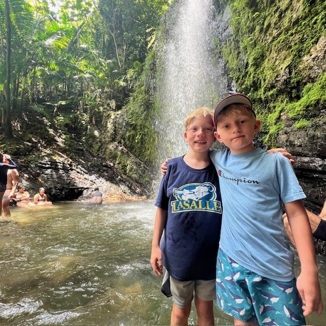 Francis & Jack stand with arms over each other's shoulders, with small waterfall behind them and other swimmers in the water beneath