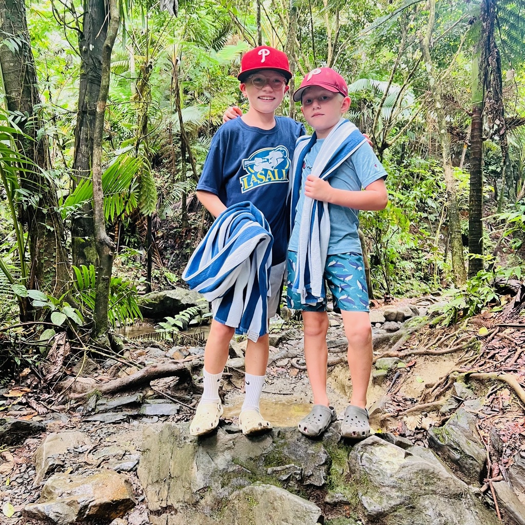Francis & Jack stand on rocks holding towels, wearing Phillies caps, among a forest of beautiful trees