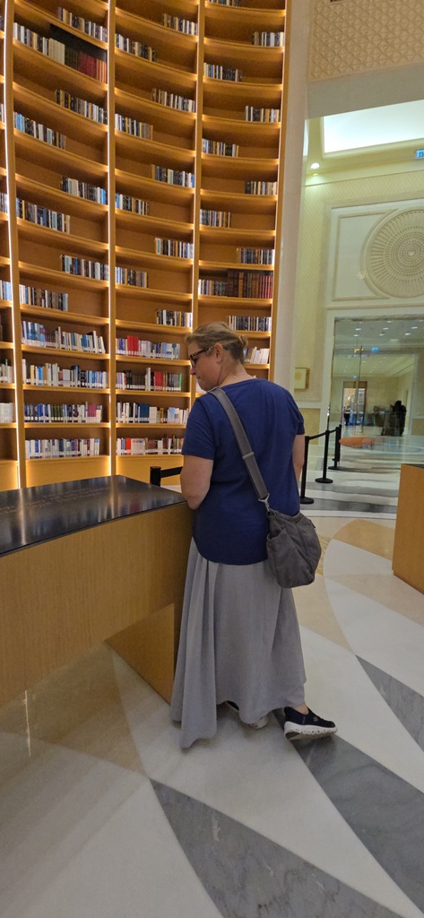 Ms. Goldberg stands in front of a huge bookshelf containing books that extends to the ceiling, with the interior of a palace in the background