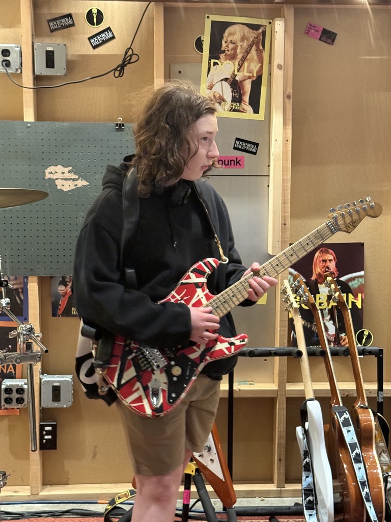 Chase stands at Roll Hall of Fame in Cleveland, OH, holding a red, white and black electric guitar, with other guitars resting in the background