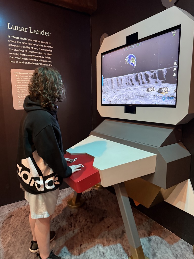Chase stands at an iteractive display of the Lunar Lander, with is hands on the controls, looking at a screen showing the moon's surface and the earth in the background