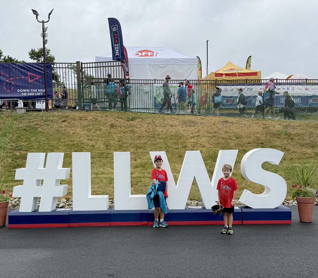 Harrison and Miles stand in front of a 3-D sign for the Little League World Series: "#LLWS", wearing red Phillies shirts, with visitors and tents behind a fence in the background