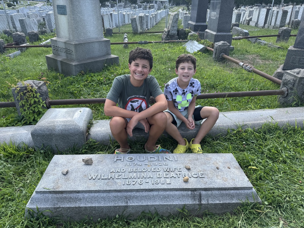 Ben & Eli sit behind the headstone that says "Houdini, 1874-1926, and beloved wife Wilhelmina Beatrice