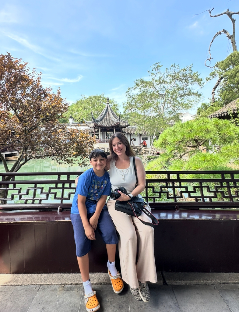 Jonathan and his mom sit on a wall with an ornamental fence in front of a pagoda with beautiful green landscape