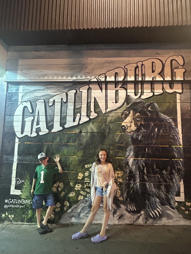 Nathaniel & Samantha stand in front of a wall with a beautiful mural of a bear and letters "GATLINBURG"