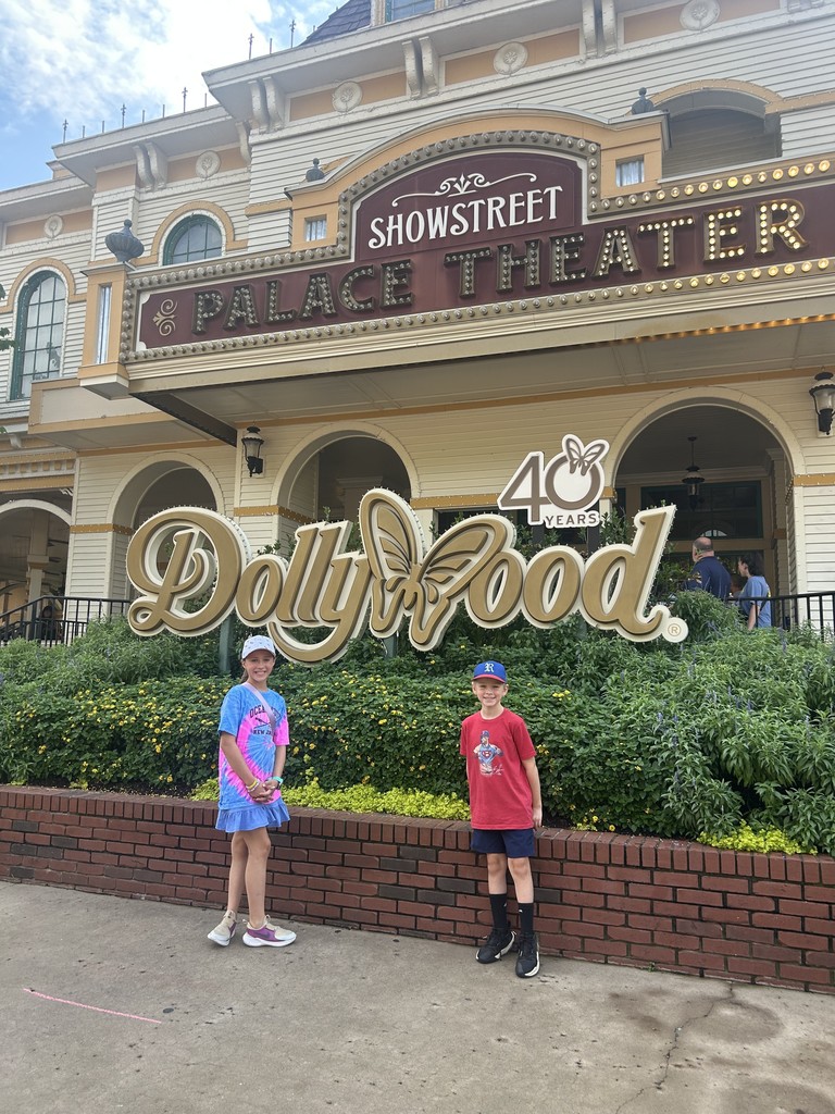 Samantha & Nathaniel stand in front of a theater with a sign "Dollywood" and "Showstreet Palace Theater", with shrubbery and a brick wall in front