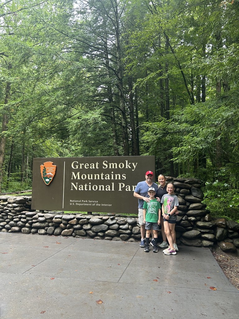 Samantha, Nathaniel and their parents stand before a sign reading "Great Smoky Mountains National Park" on a round stone wall, with beautiful forest behind