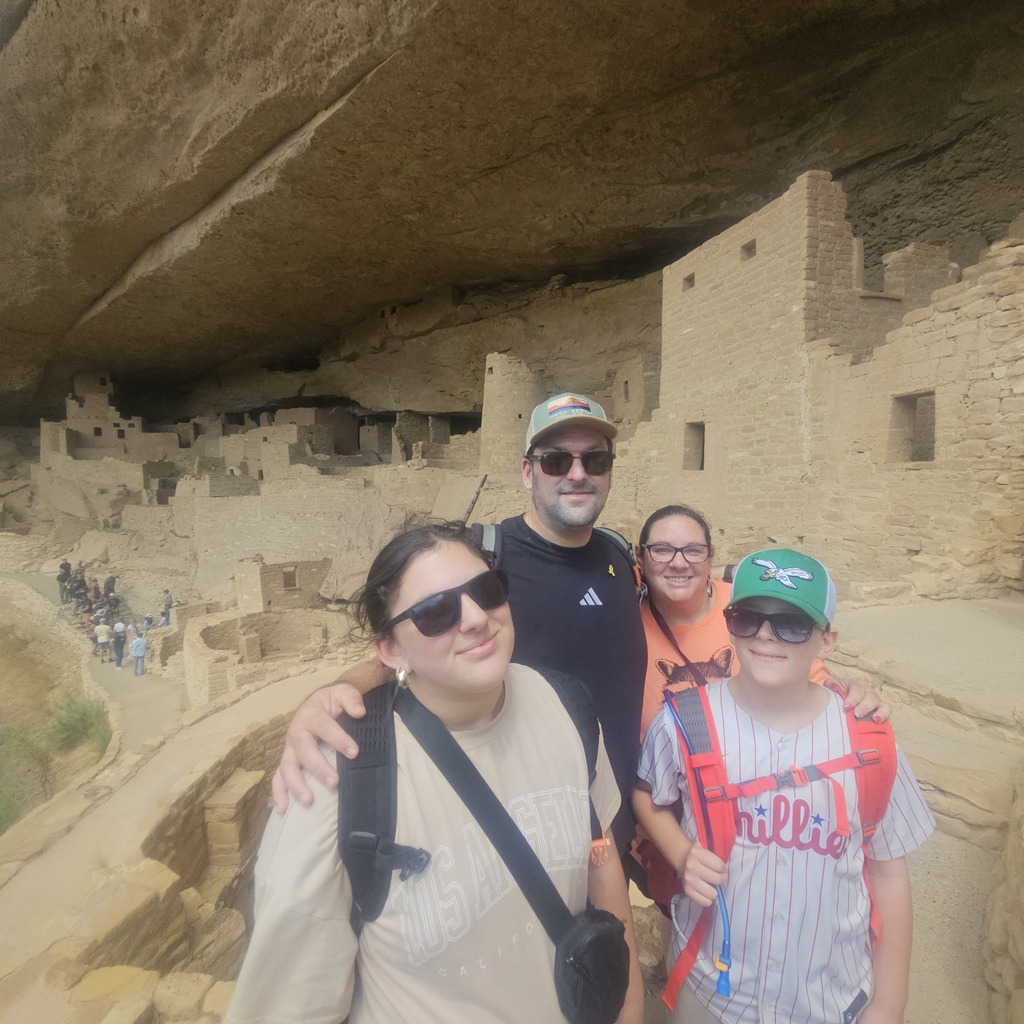 Adina, Ben and parents stand in front of an adobe house buildi out of sand with tourists in background