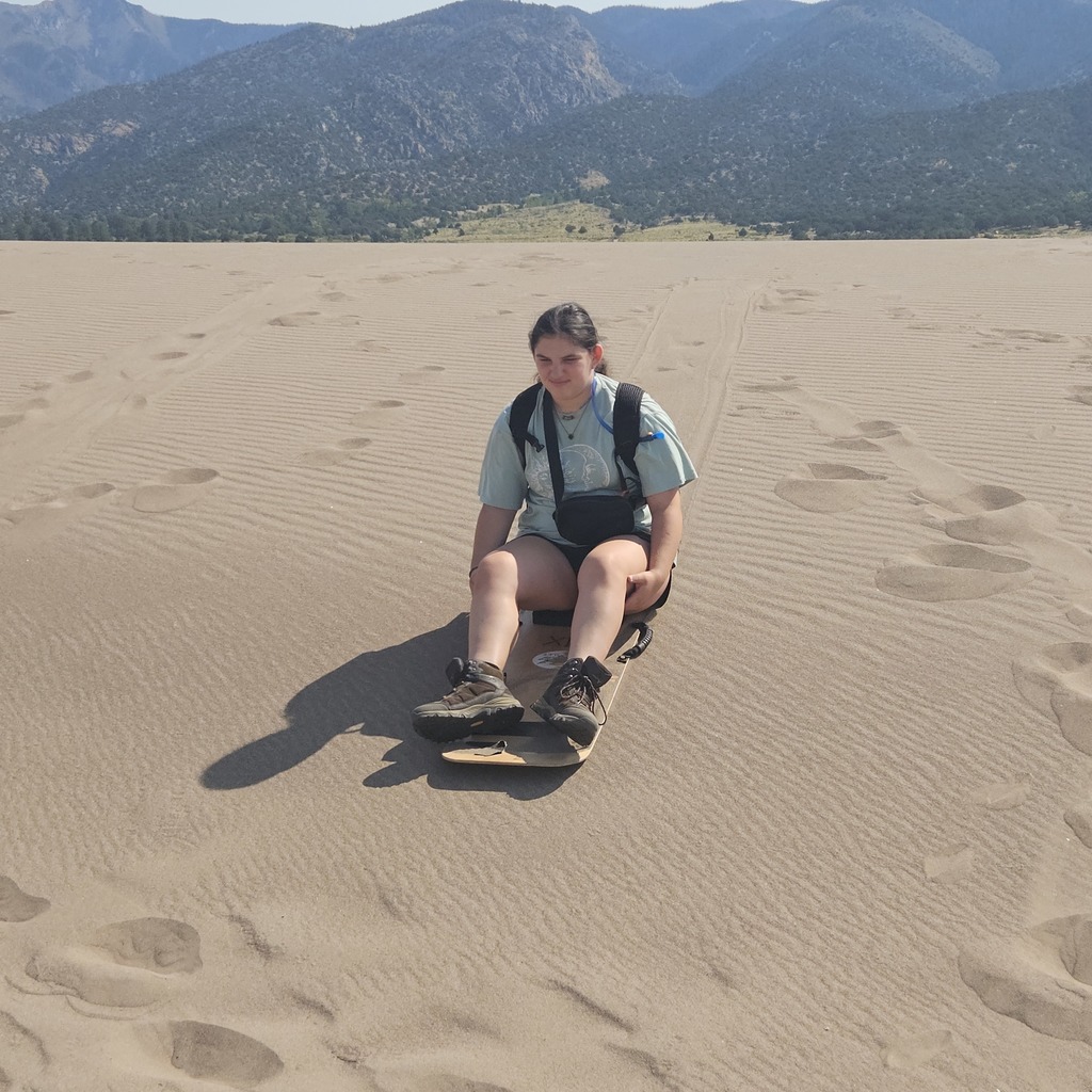 Adina rides a sand board on the sand, with mountains in background