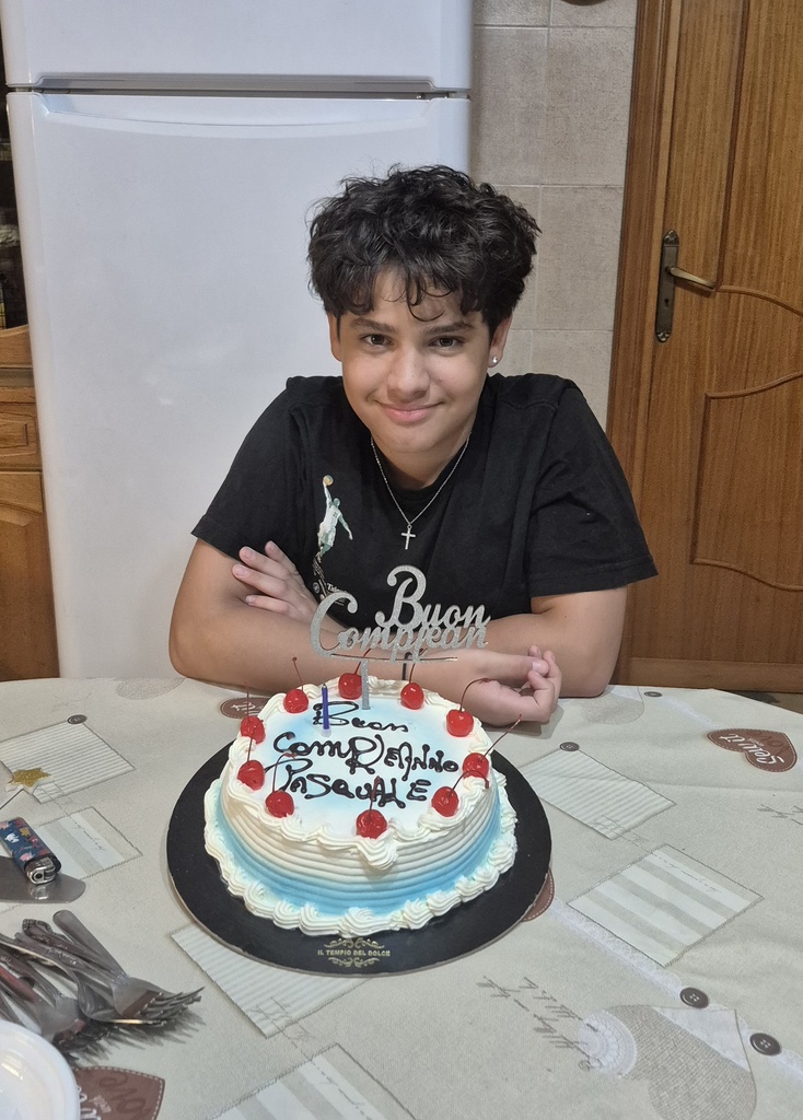 Pasquale sits at a table smile with his birthday cake in front of him, which has blue shadors, whipped cream and cherries