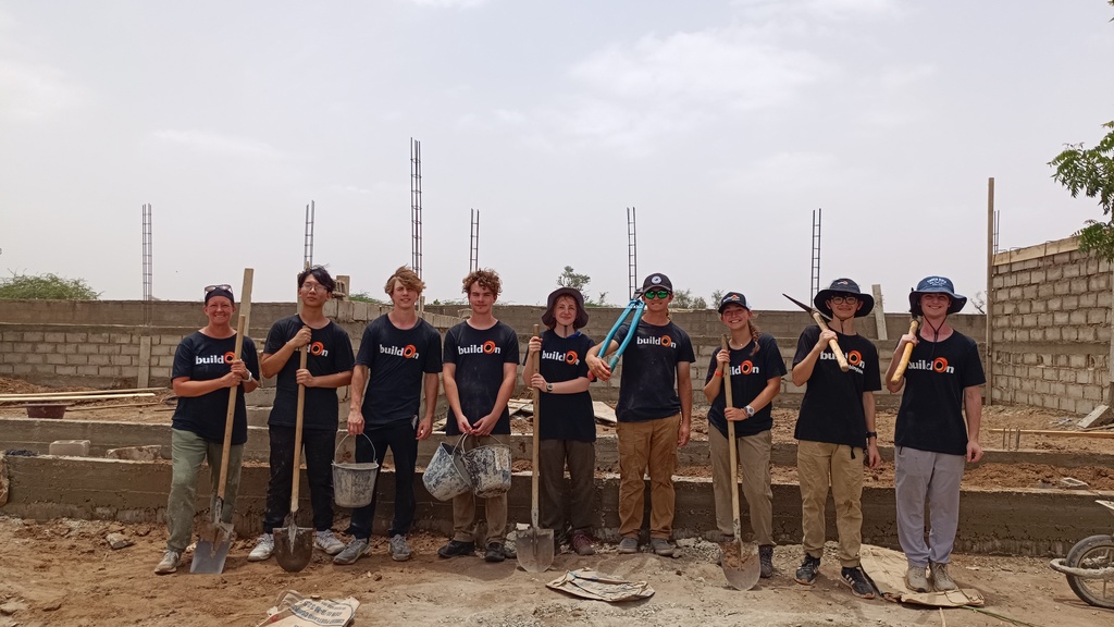 9 students in black t-shirts stand in front of a building they are helping to construct
