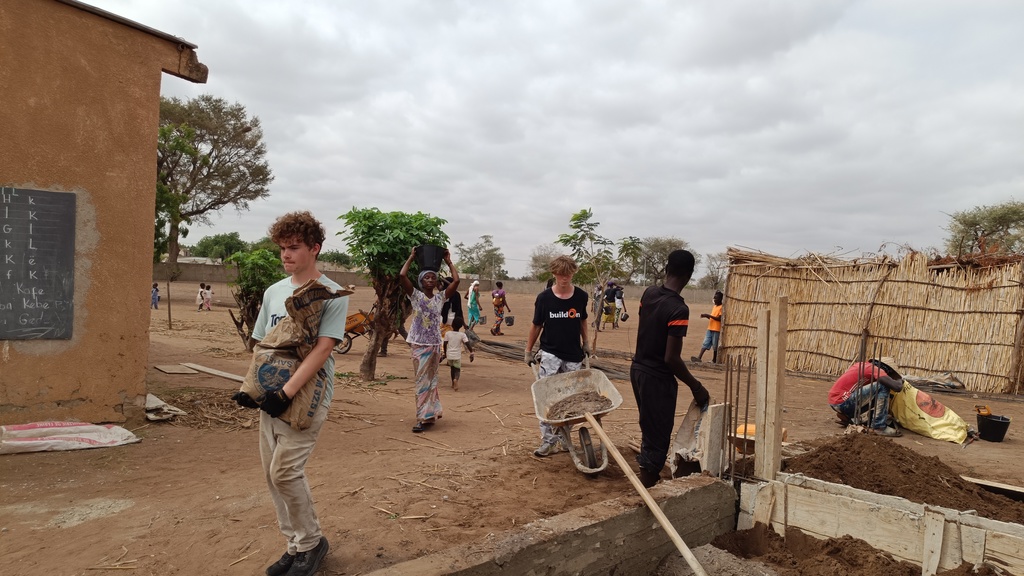Students work together with community members by pushing a wheelbarrow and carrying building supplies