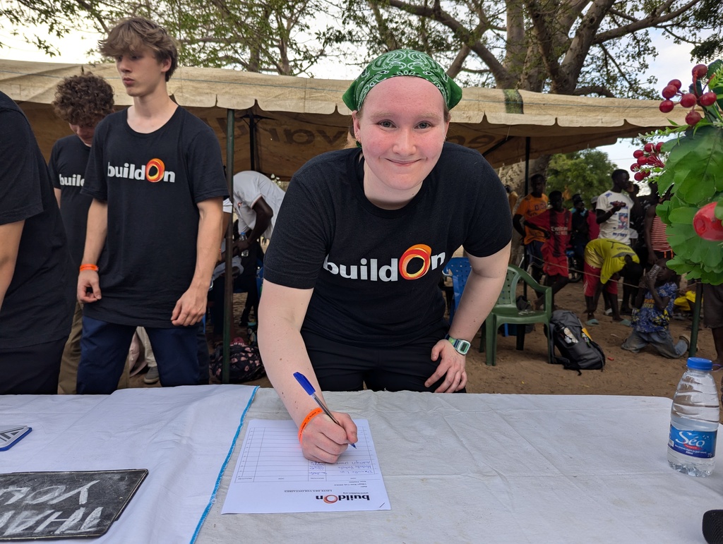 A student wearing a green bandana and black t-shirt leans forward over a talble to write, with other students and community members in the background