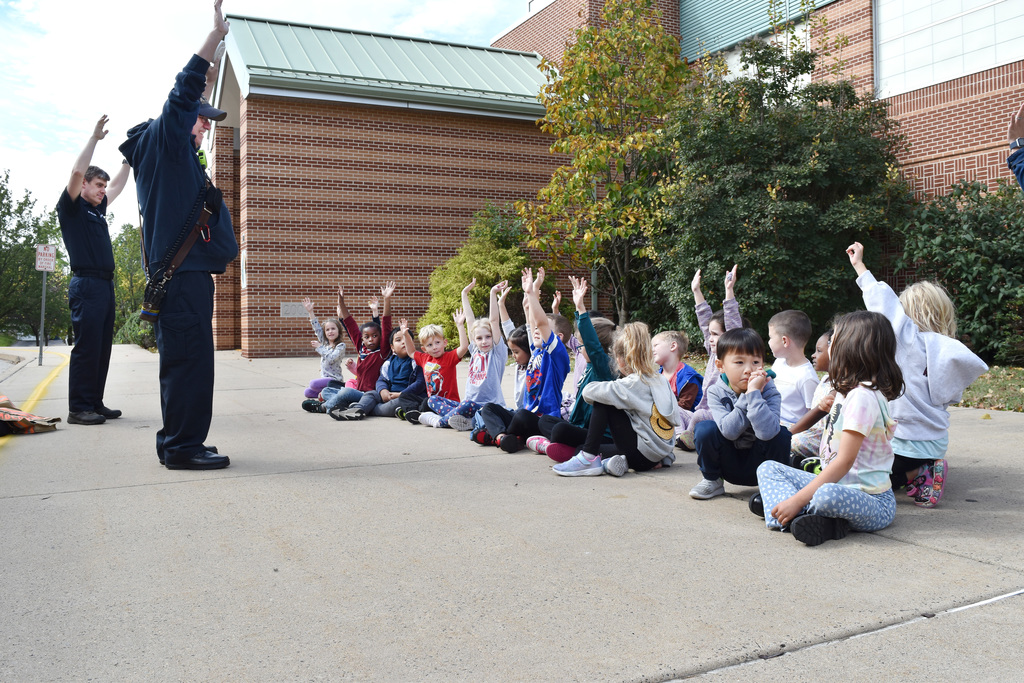Firefighters stand with hands raised in the air, along with kindergarteners sitting on the sidewalk during a discussion of fire safety