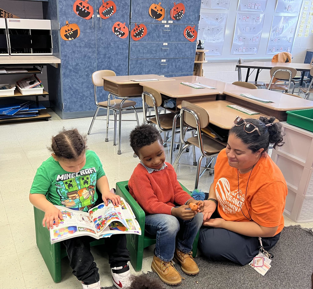 Mrs. Oldham sits on the floor next to 2 students, one of whom is looking at a book