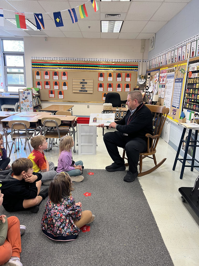 Dr. Fecher sits in the front of the classroom reading to students, who sit on a carpet before him, listening