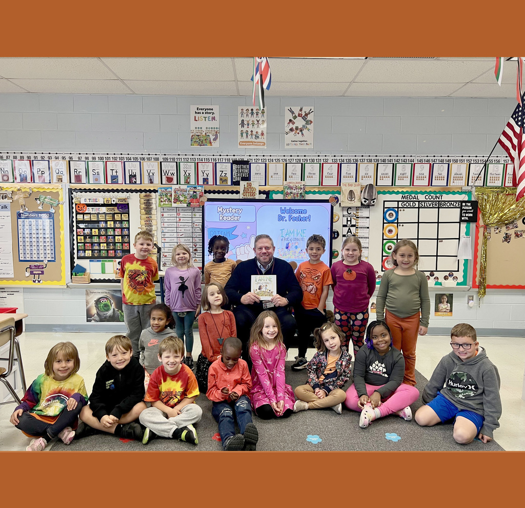 Dr. Fecher sits in front of classroom holding the book "I am We: A Book of Community" with the class of first graders standaround him