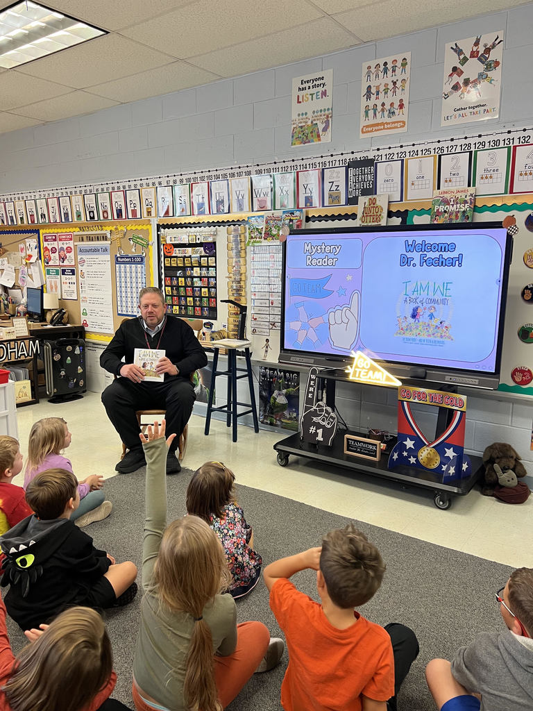 Dr. Fecher sits in a chair speaking to students, some of whom have raised hands, with a screen reading "WElcome Dr. Fecher!" behind him