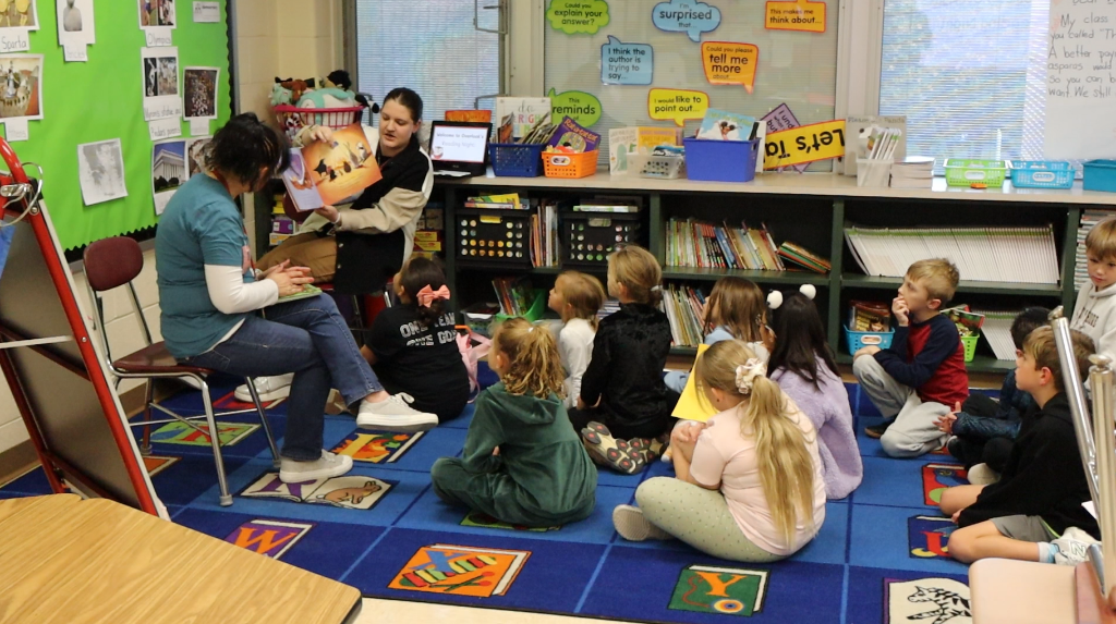 Teachers read to students who are sitting on a carpet in front of them