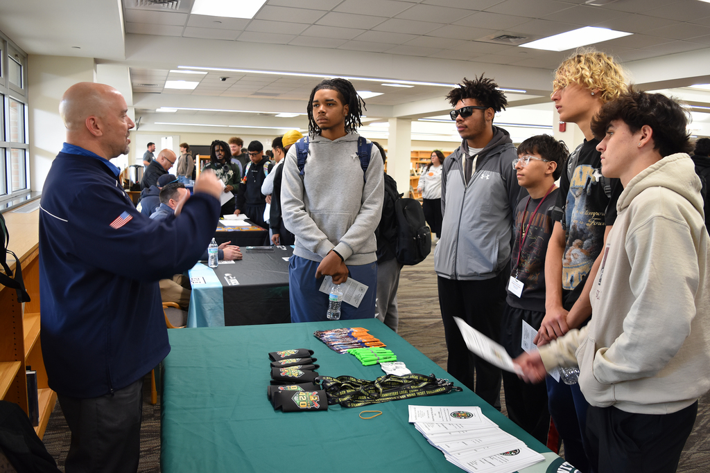 Students are gathered around a table with literature, listening to a representative answering questions