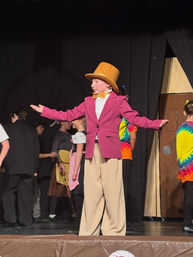 A student dressed on pink suit jacket, tan trousers and a tan top hat performs on stage