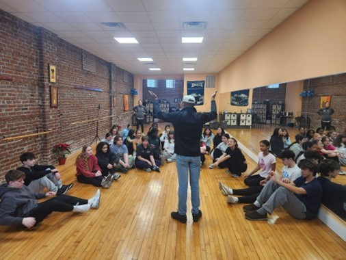 Students sit on either wall of a dance studio, listening to the instructor in the middle