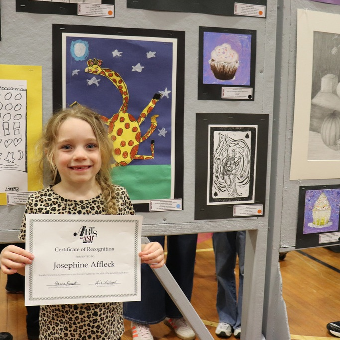 A young student stands in front of a display of artwork, holding her Certificate of Recognition