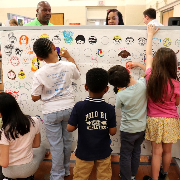 Seven students stand in front of a vertical drawing board and add their own personal artwork