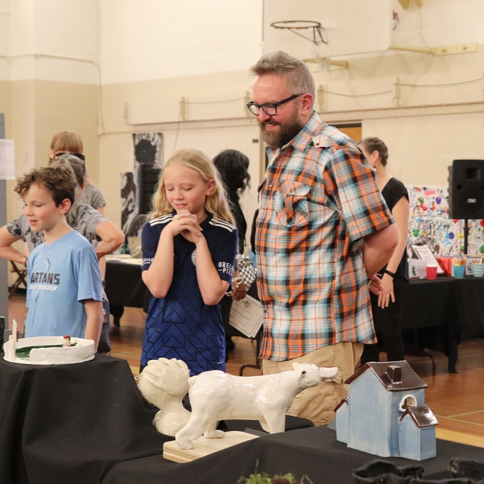 Several students and a parent smile as they admire the 3-D artwork on display tables
