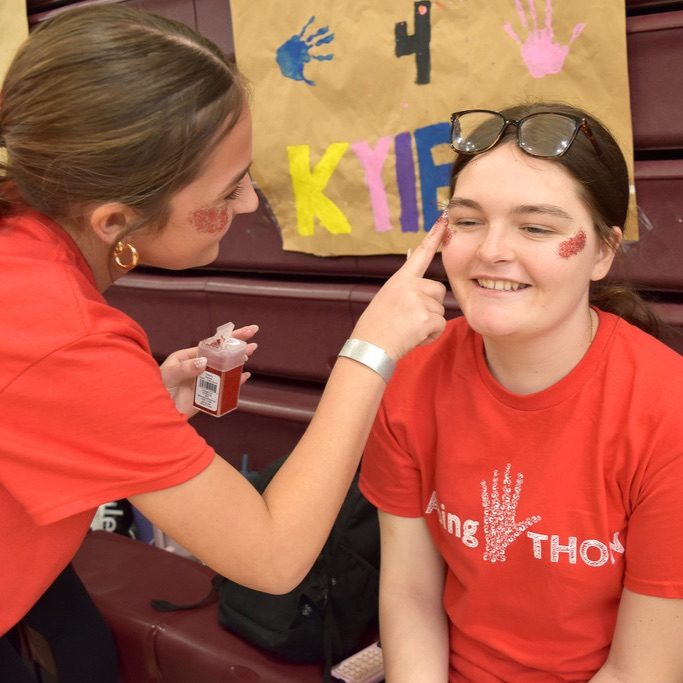 A student applies red glitter makeup to another student's face during AbingTHON