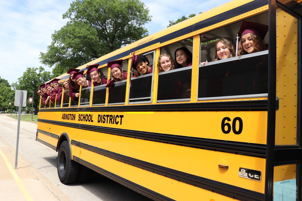 Students hang out of school bus windows in their caps and gowns