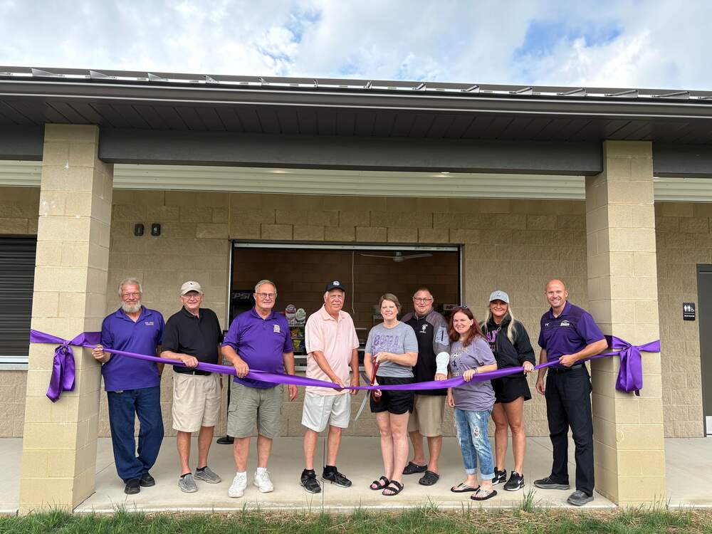 Nine people standing in front of a cinder block building holding a purple ribbon with large scissors 