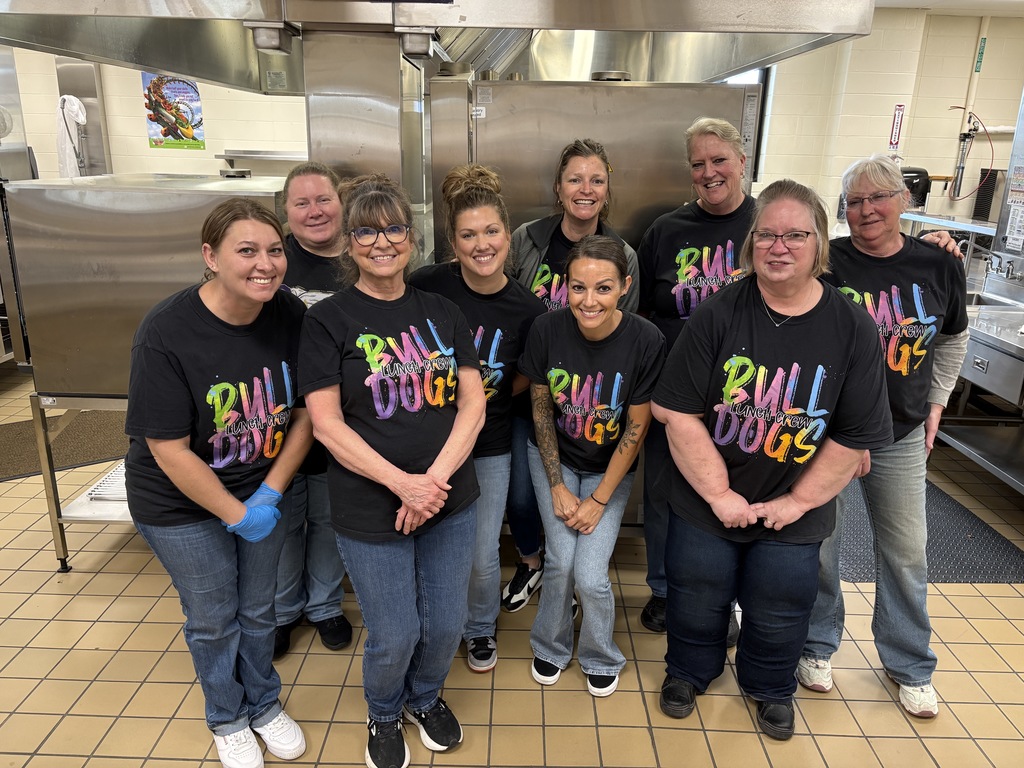 Nine women in black shirts with tie dye text standing in front of the cafeteria 