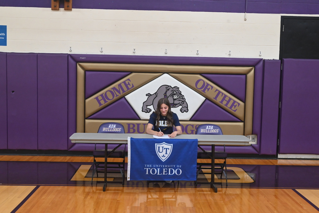Student sitting at table with purple and gold bulldog background and college flag