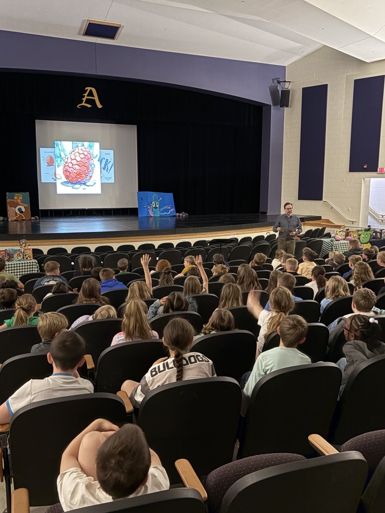 Adult standing in front of students seated in an auditorium reading a book