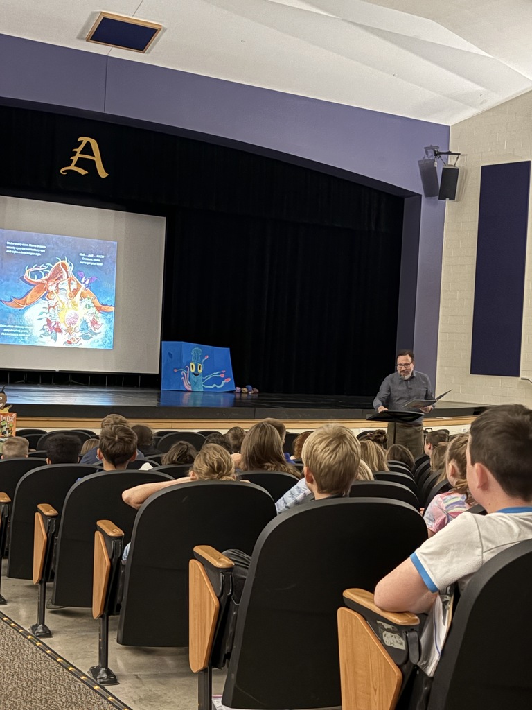 Adult standing in front of students seated in an auditorium reading a book