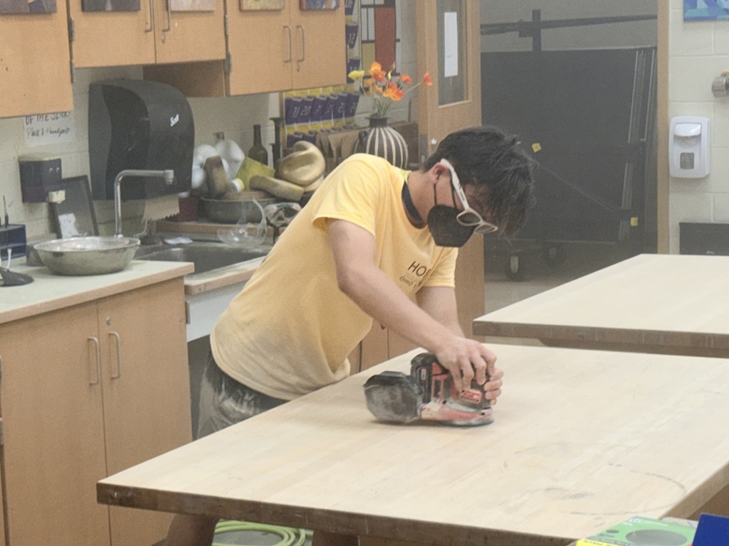student in yellow shirt sanding tables with electric sander