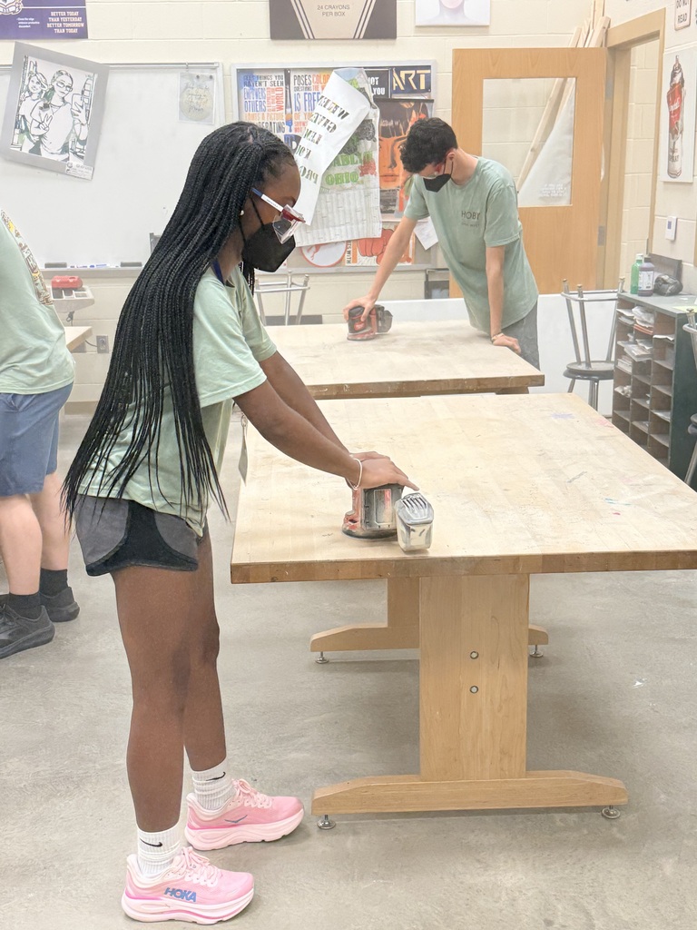 student in yellow shirt sanding tables with electric sander