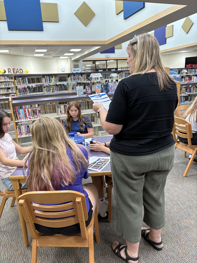 An adult standing at a table holding a piece of paper talking to studne