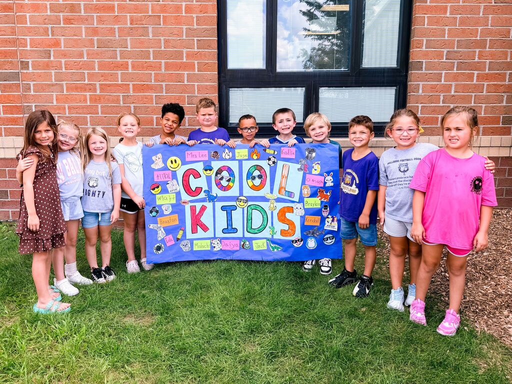 Twelve students holding a paper sign that spells out the name Cool Kids in front of a brick building