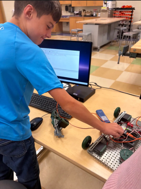 Student standing at the desk in front of a computer with his hand on a robot